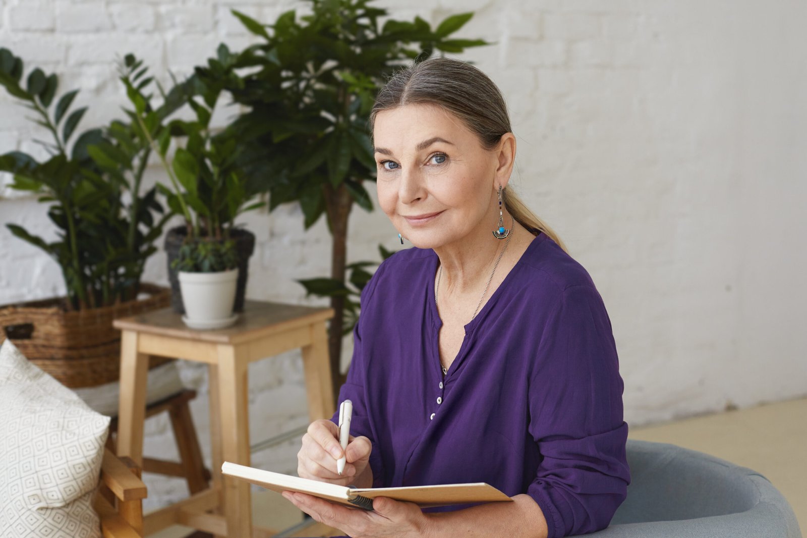Portrait of busy modern 50 yeard old middle aged woman in violet shirt writing in copybook, making plans, looking at camera with positive friendly smile, sitting on chair, surrounded with green plants